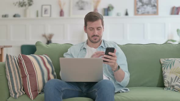 Young Man with Laptop Using Smartphone on Sofa