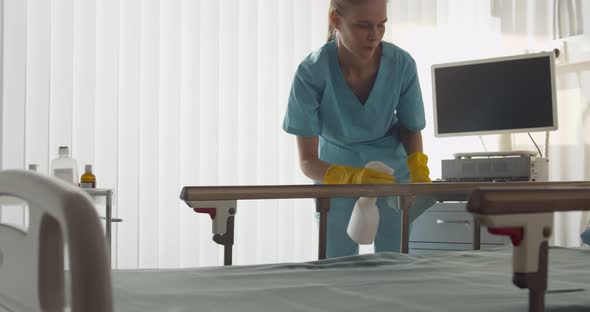 Young Nurse in Scrubs Cleaning Bed with Detergent Sprayer in Hospital Ward