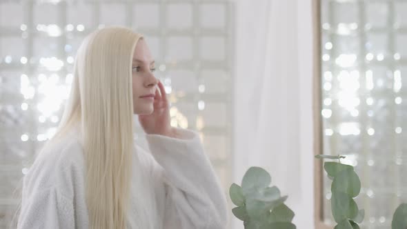 Portrait of Attractive Young Blond Woman Wearing Bathrobe With Natural Makeup Looking in Mirror