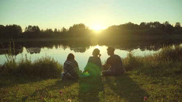 Young People Enjoy Vacation Sitting on Riverbank at Sunset