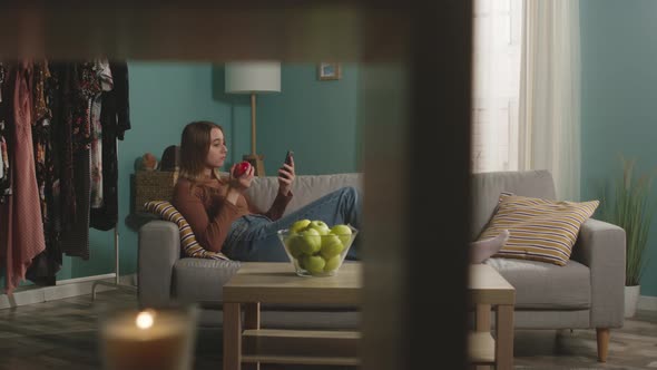 Young Woman with Red Apple Is Lying on Sofa and and Watching Something Gadget