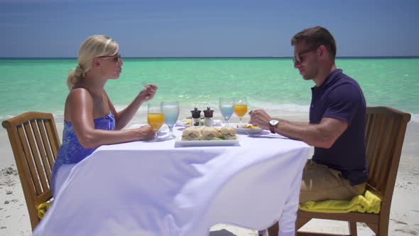 A man and woman eat breakfast on a tropical island beach