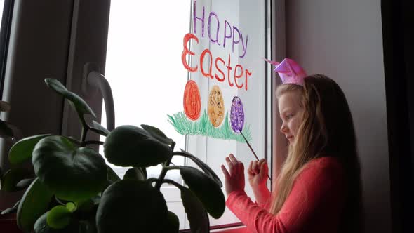 Cute little girl in bunny ears painting Easter eggs with green spring grass on window at home