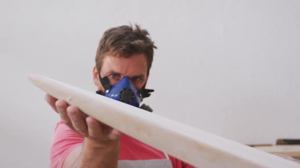 Caucasian male surfboard maker wearing a breathing face mask and checking a wooden surfboard