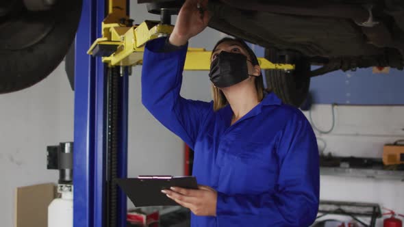 Female mechanic wearing face mask taking notes on clipboard under a car at a car service station