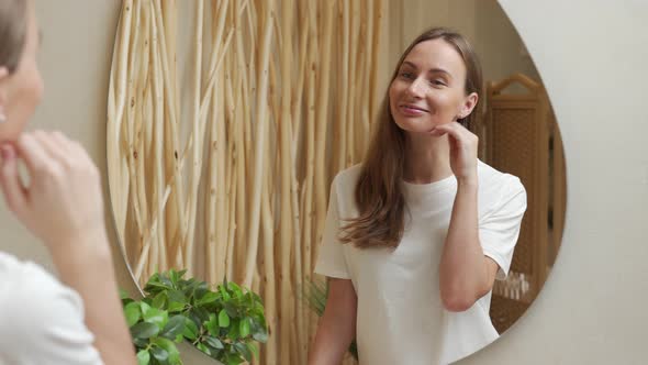 Young Woman Touches Her Face and Smiles While Looking in the Mirror While Standing in the Bathroom