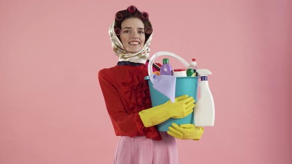 A Woman with a Headscarf and Curlers is Holding a Bucket of Cleaning Products