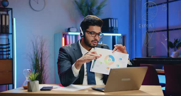 Office Manager in Suit Wears Headset During Video Conference with Coworkers and Explaining Report