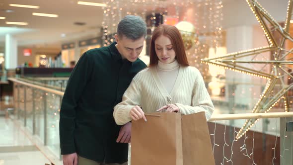 Happy Attractive Young Woman Showing Purchases in Shopping Bags to Handsome Boyfriend Standing in