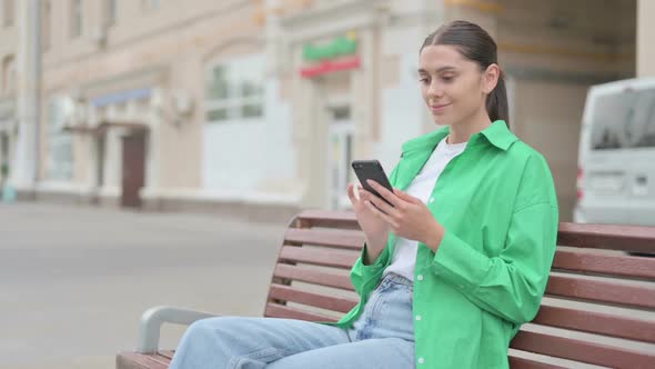 Hispanic Woman Celebrating Online Success on Smartphone While Sitting Outdoor on Bench