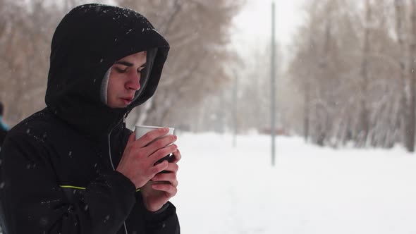 Man Stands in the Park Under Snowfall and Drinks Hot Drink