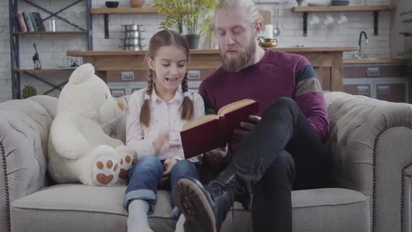 Handsome Caucasian Man Turning Book Pages and Talking To Teen Girl. Daughter Laughing As Listening