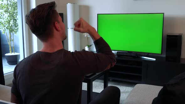 A Man Watches a TV with a Green Screen in a Cozy Living Room and Begins To Celebrate