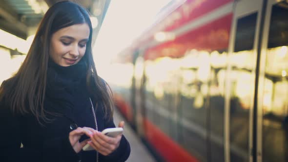 Young Lady Typing on Smartphone on Platform Near Train and Smiling to Camera
