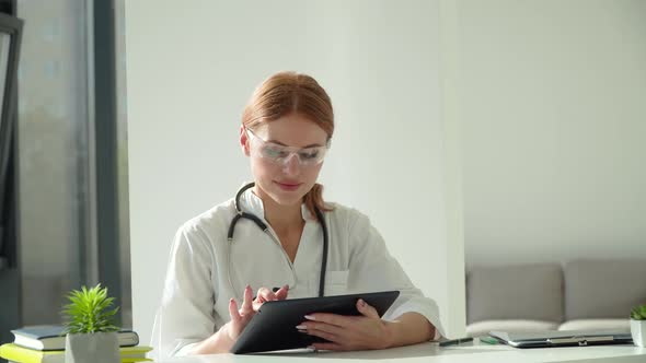 Female Doctor in White Coat, Protective Glasses and Stethoscope Over Her Neck Using Tablet at Desk