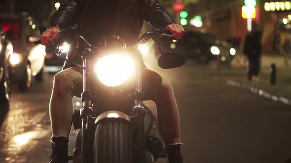 Front View of a Bearded Motorcyclist Drives a Bike in the City with Bright Headlight at Night