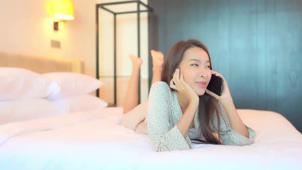 Smiling Young Asian Woman Talking on Smartphone With Friend or Lover in Privacy of Bedroom, Lying on