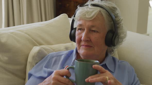 Caucasian senior woman wearing headphones listening to music while holding coffee cup at home