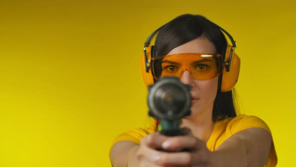 Brunette Woman in Protective Headphones and Glasses is Holding a Drill Like Gun