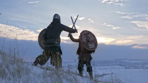 Two viking are fighting with axes and shields on the winter meadow.