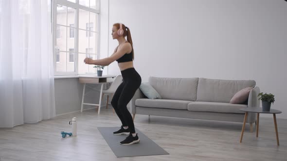 Athletic Woman Doing Jump Squats Exercising Wearing Headphones At Home