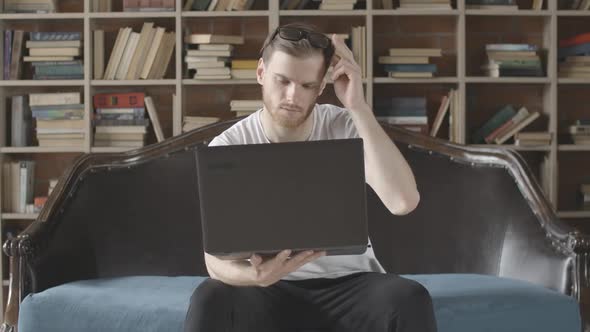 Absorbed Young Man Sitting with Laptop at the Background of Bookshelves and Looking at Screen