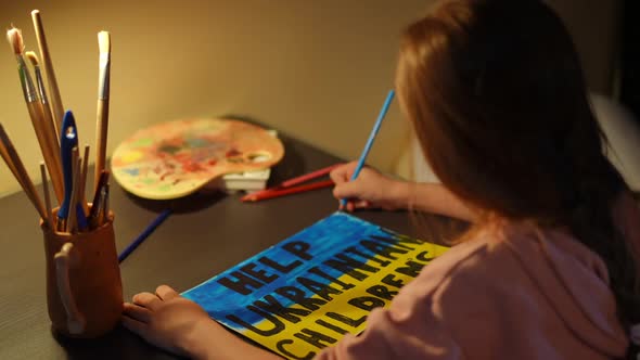 Shooting Over Shoulder of Cute Little Girl Painting Help Ukrainian Children Placard Sitting at Table
