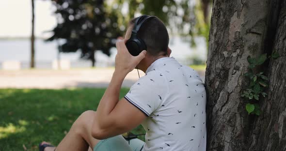 Man Puts on Big Headphones Leaning on Tree