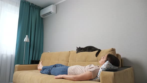 Young Woman and Cat Lie on a Sofa in a Room with an Air Conditioner