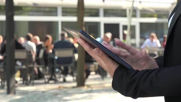 A Businesswoman Works on a Tablet in a Street in an Urban Area - People Sit Outside a Restaurant