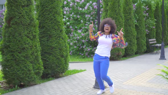 Cheerful Happy African Woman with an Afro Hairstyle with Headphones Walking Down the Street Dancing