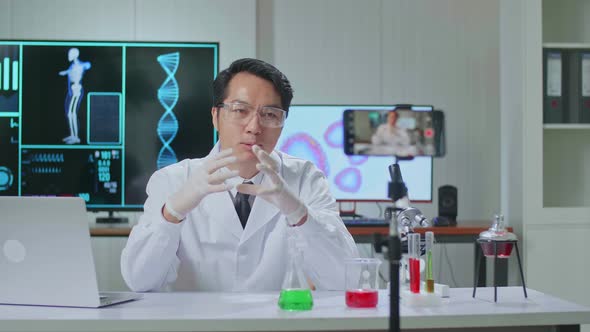 Asian Scientist Man Recording Webinar, Using Smartphone On Tripod, Sitting At Desk In Office