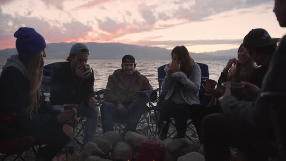 Group of friends drinking from mugs around campfire during sunset