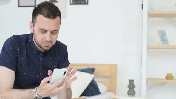 A Young Man in a Blue Tshirt is Sitting on a Sofa and Looking for Information on the Phone