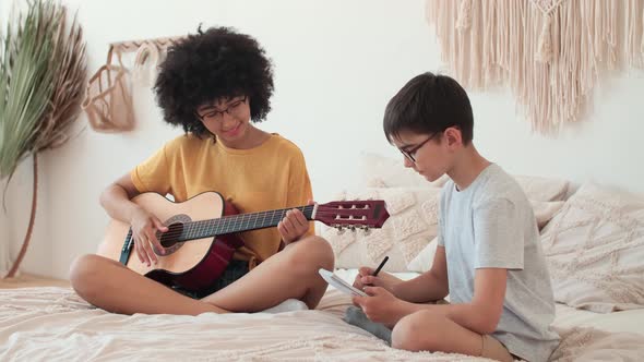 Afro American Girl Teaches Boy to Play Guitar While Sitting on Bed at Home