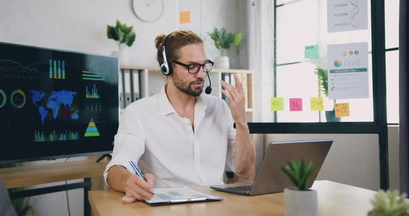 Man in Glasses wears Headset Holding Video Meeting on Laptop from Office