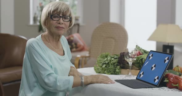 Blond Charming Caucasian Senior Woman in Eyeglasses Sitting at Table with Blue Screen Laptop Looking