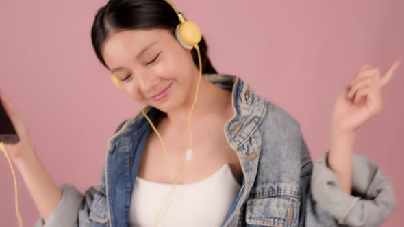 Happy pretty Asian teenager girl dancing and listening music by smartphone on pink background.