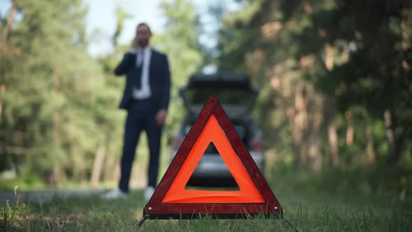 Red Triangle Emergency Road Sign on Green Grass with Blurred Young Caucasian Man Talking on Phone at