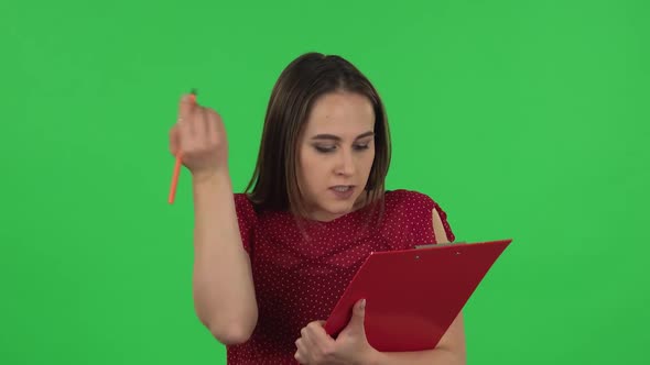 Portrait of Tender Girl in Red Dress Fills Papers in Red Folder with Pensil. Green Screen