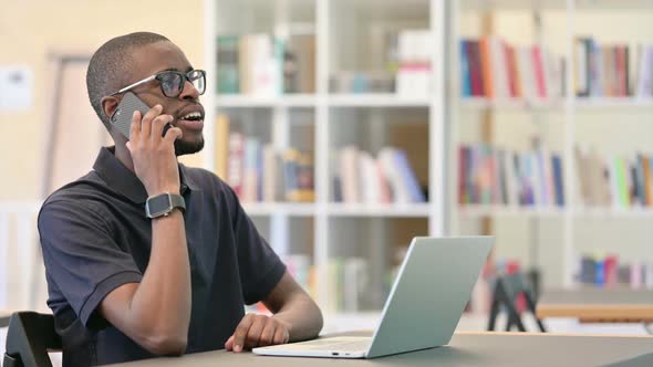 Young African Man with Laptop Talking on Smartphone in Library