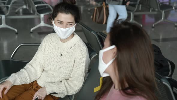 Two Women Friends Tourists in Medical Mask Sitting in Airport Terminal at Safe Distance and Talking