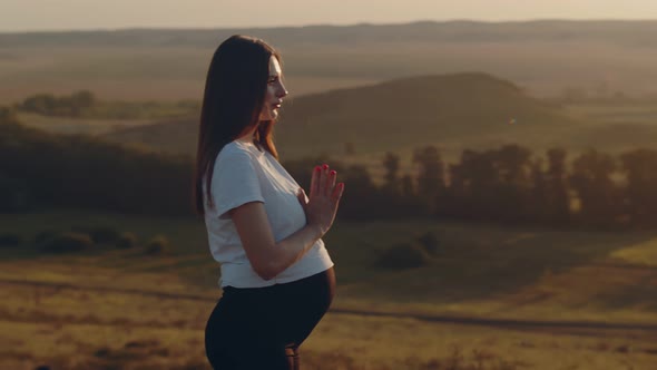 Pregnant White Woman Stands Outdoor and Meditates with Her Hands Folded to Each Other