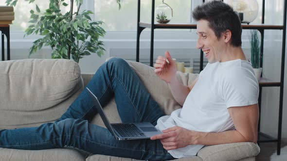 Caucasian Man Millennial Adult Guy Lies Sitting on Couch Wears Jeans and White Shirt Uses Laptop for