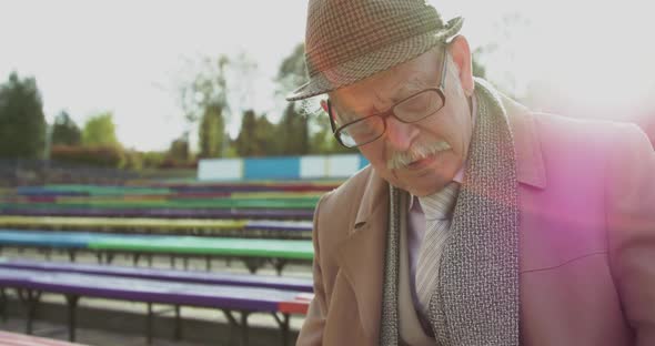 Portrait of Serious Grandfather Reading Newspaper on Bench in Green Sunny Park