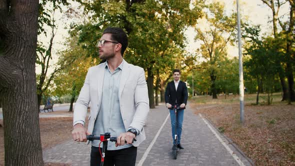 Portraits of Two Handsome Young Businessman Riding an Electric Scooters on the Road in City Park