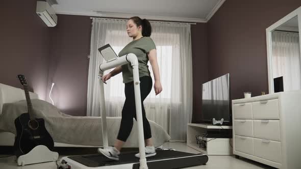 Young chubby woman is jogging on treadmill at home