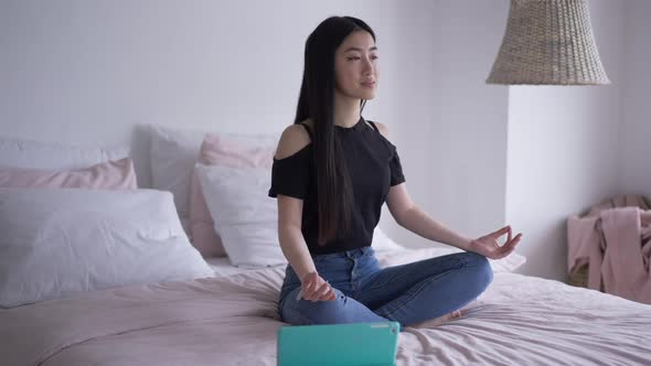 Wide Shot of Calm Confident Asian Woman Sitting in Lotus Pose on Bed Smiling with Tablet Lying Aside