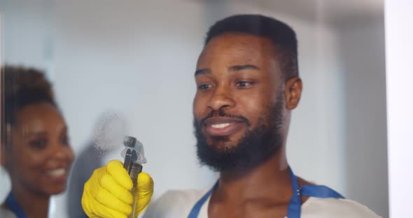 African Male Janitor Using Sprayer and Rag To Clean Window in Office Wearing Apron and Gloves