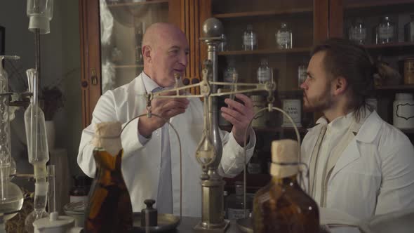 Happy Old Caucasian Man Showing Vintage Test Tube and Flask To Young Man and Talking. Apothecaries